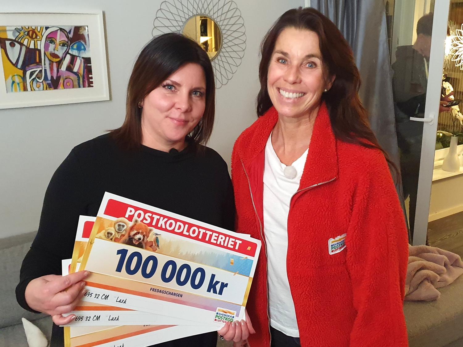 Två kvinnor står inomhus och ler; den ena håller stora lottericheckar, bland annat en på 100.000 kr, med 