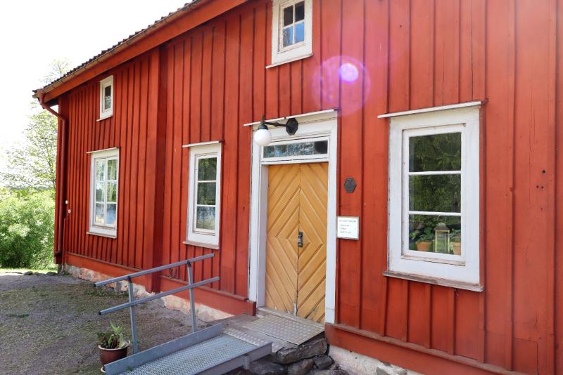 En röd träbyggnad med vitmålade fönster, en lampa ovanför huvuddörren, en metallramp som leder till entrén och en liten plakett bredvid dörren.