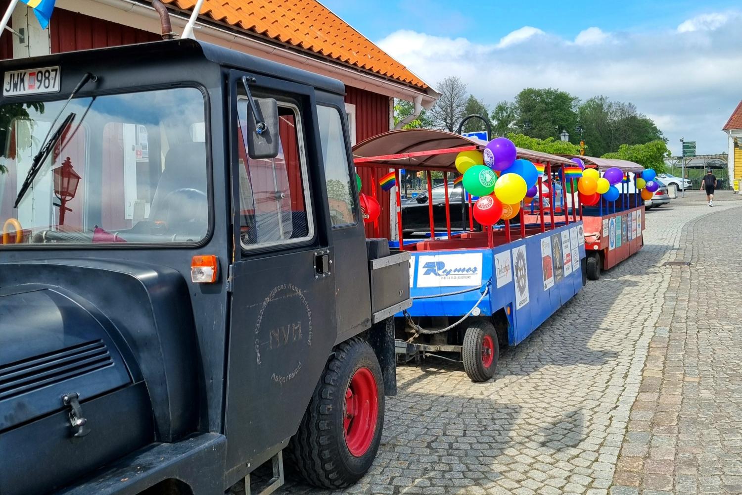 Ett litet svart tåg med färgglada personvagnar dekorerade med ballonger står parkerat på en kullerstensgata nära röda träbyggnader.