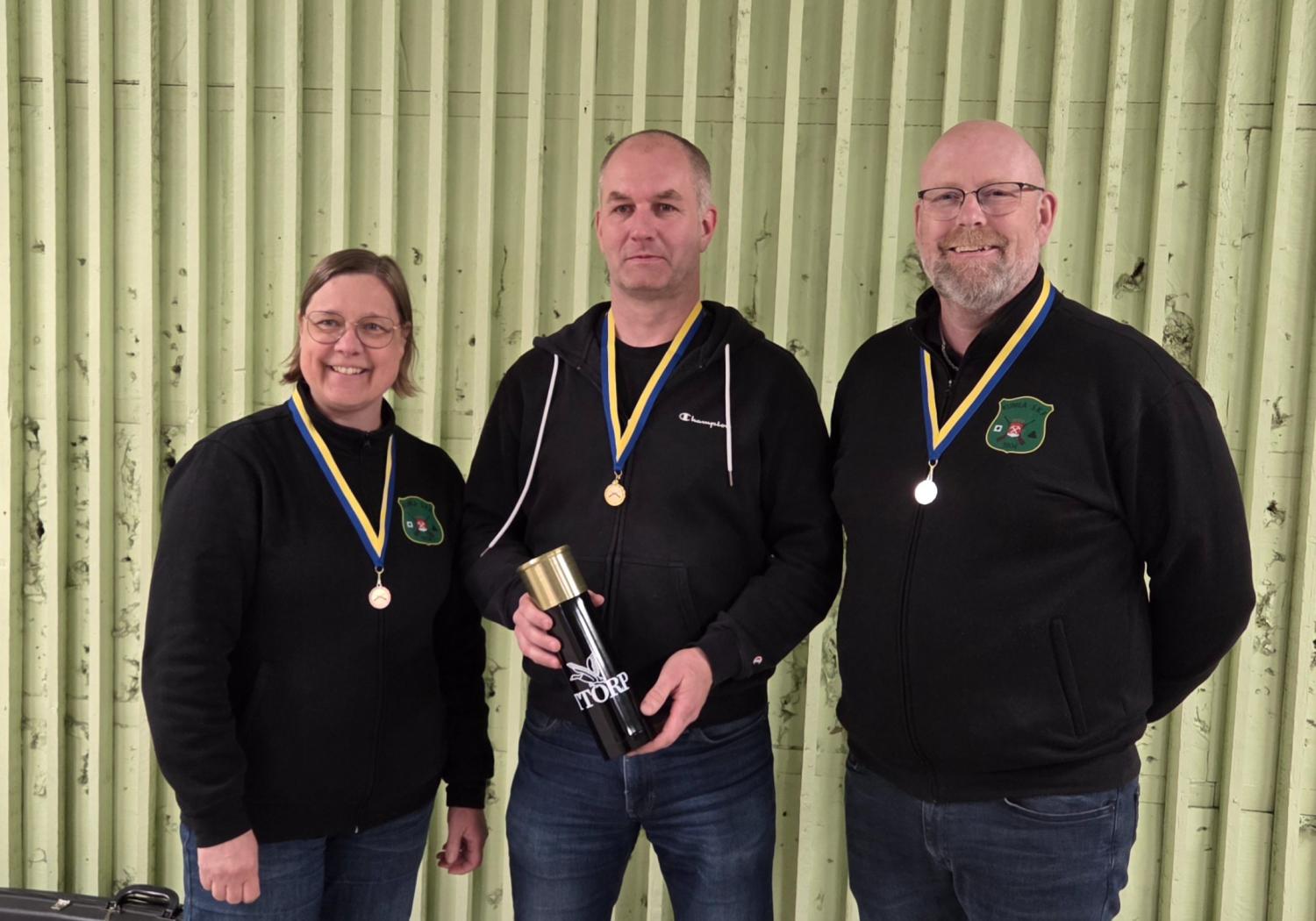 Tre medaljförsedda personer står framför en ljusgrön vägg. Personen i mitten håller i en termos i svart och guld. Alla är klädda i mörka kavajer.