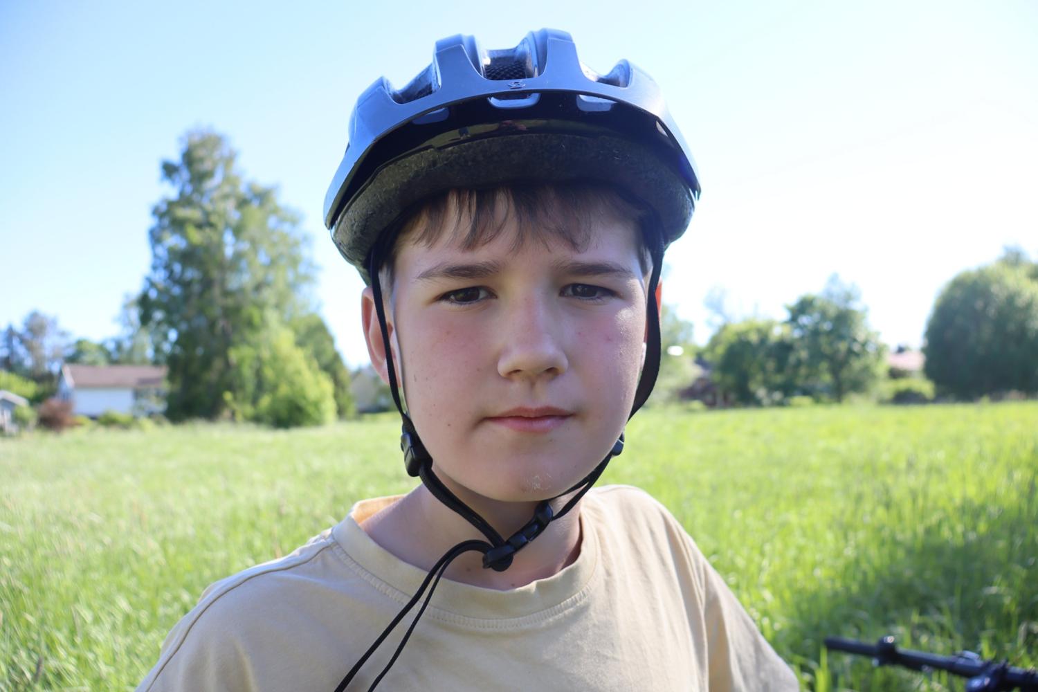 En pojke med cykelhjälm står utomhus på en gräsplätt en solig dag och tittar rakt in i kameran.