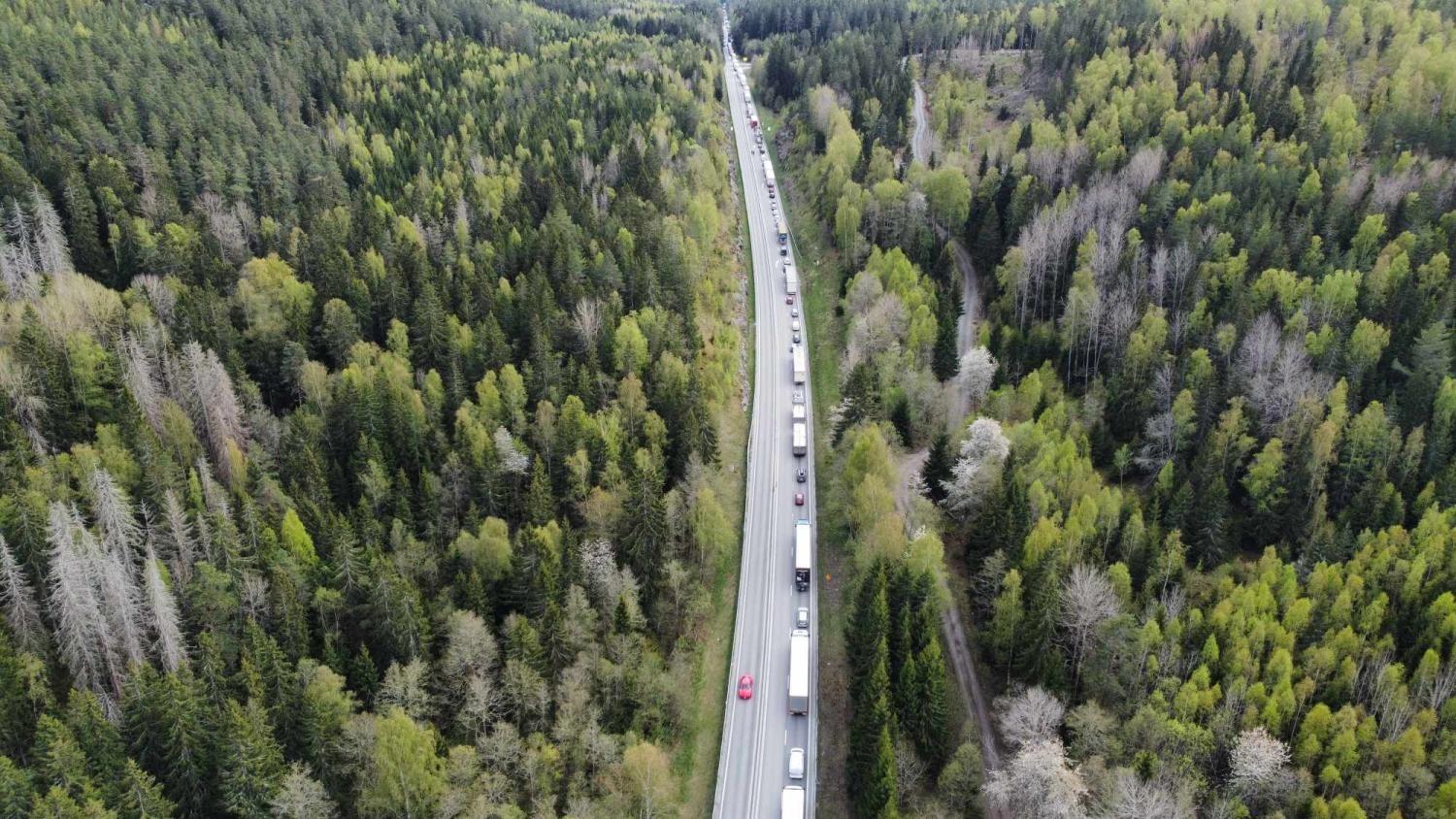 En lång rad fordon, främst lastbilar, sitter fast i trafiken på en tvåfilig väg omgiven av tät grön skog.