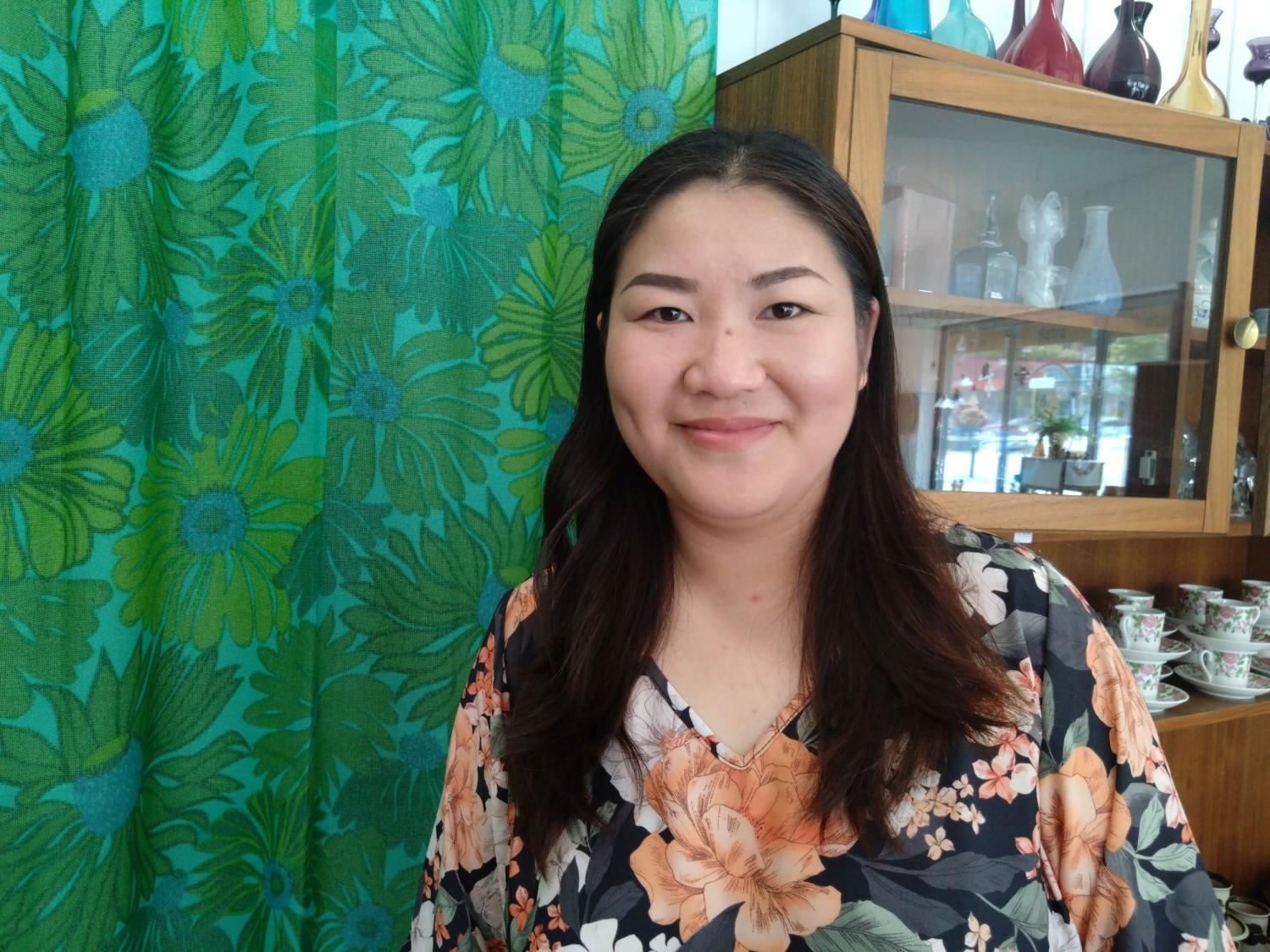 En kvinna i en blommig topp står framför ett träskåp med glas och en grön blommönstrad gardin.