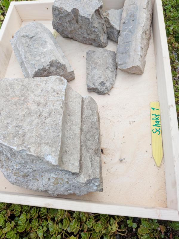 En träbricka med flera grova stenfragment märkta ”Schlack 1”, placerad på en grön underläggsmattor.