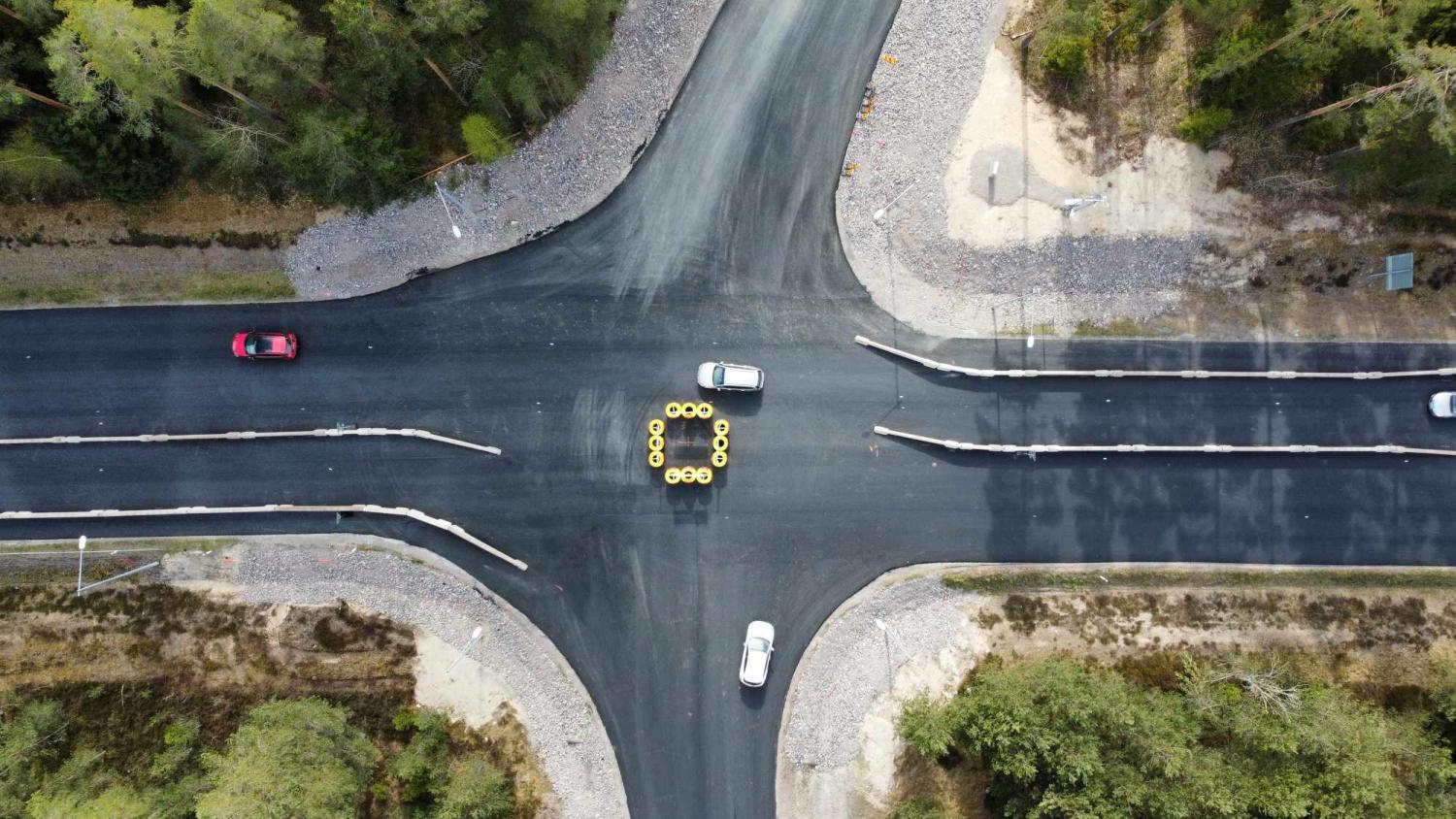 Flygfoto över en fyrvägskorsning med tre bilar och ett kluster av gula tunnor arrangerade i en cirkel i mitten. Omgivningen är skogsklädd.