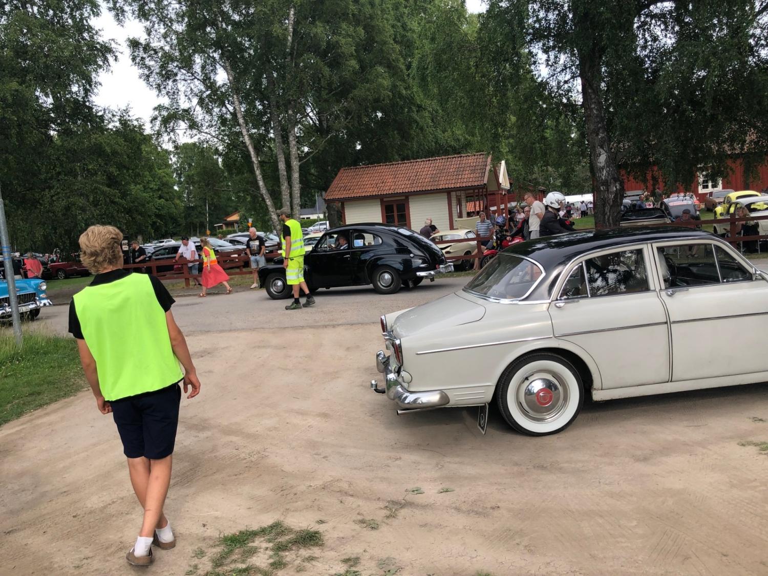 En grupp människor i neonvästar dirigerar veteranbilar under ett utomhusevenemang, med träd och byggnader i bakgrunden.