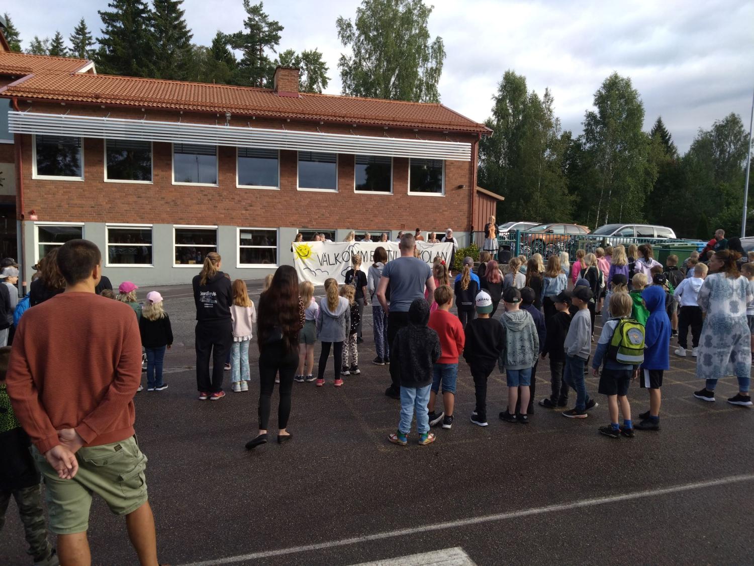 En skara människor, inklusive barn, är samlade utomhus nära en tegelbyggnad med en banderoll. Vissa är vända mot byggnaden medan andra står i grupper. Träden syns i bakgrunden.