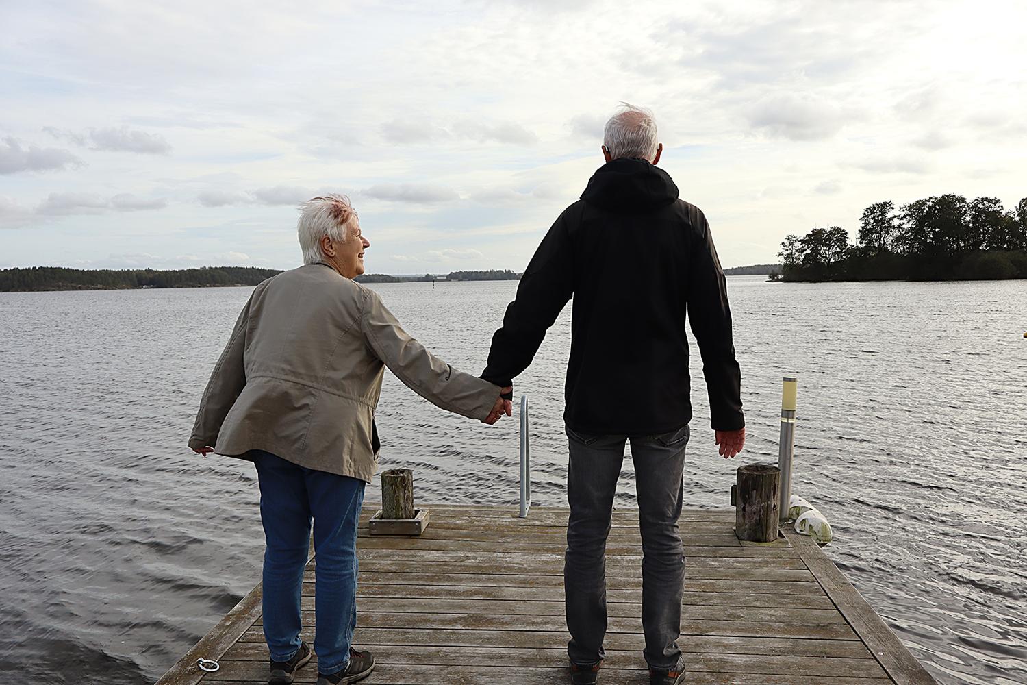 Två äldre vuxna står på en träbrygga med utsikt över en sjö och håller varandra i handen. Himlen är molnig och träd syns i fjärran.