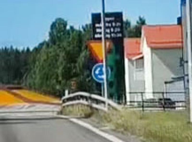 En väg med ett metallräcke på vänster sida, en blå rondellskylt och en digital väginformationsskylt nära hus med orange tak. I bakgrunden står träd på rad.