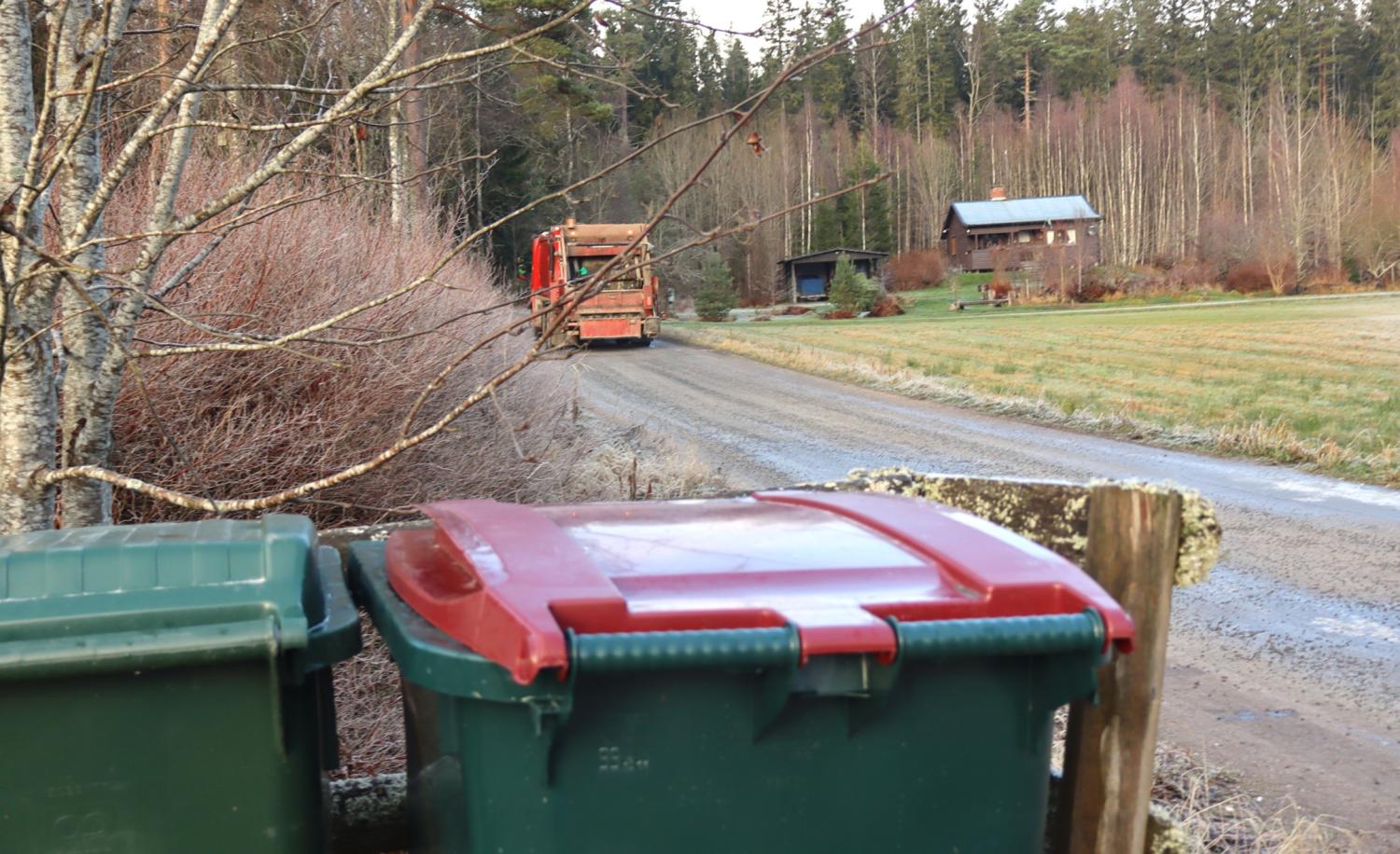 Två soptunnor står i förgrunden nära en grusväg, en lastbil kör iväg och i bakgrunden syns hus omgivna av träd.