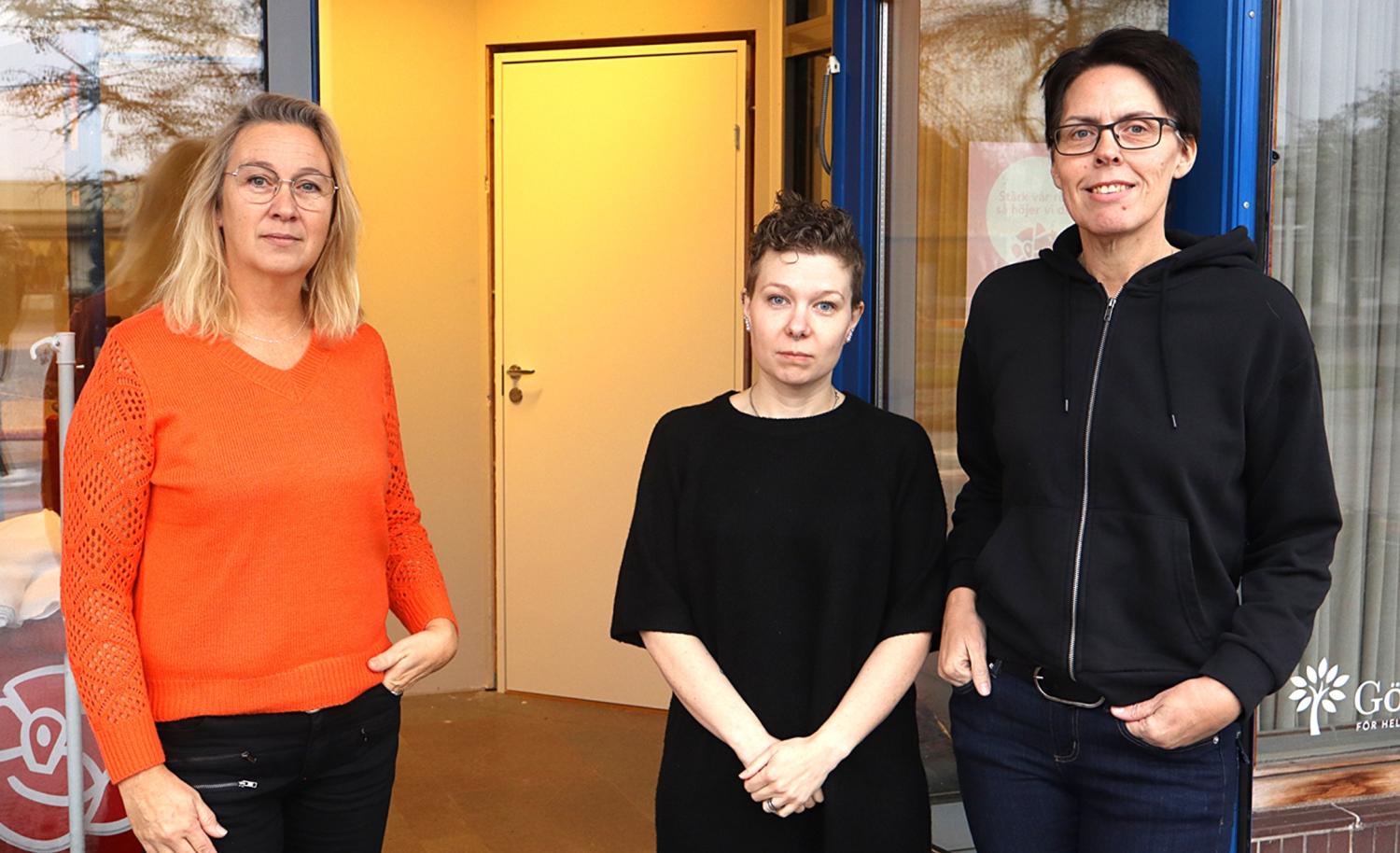 Tre personer står utanför en entré till en byggnad och poserar för fotografering. Två bär glasögon och alla är klädda i vardagskläder.