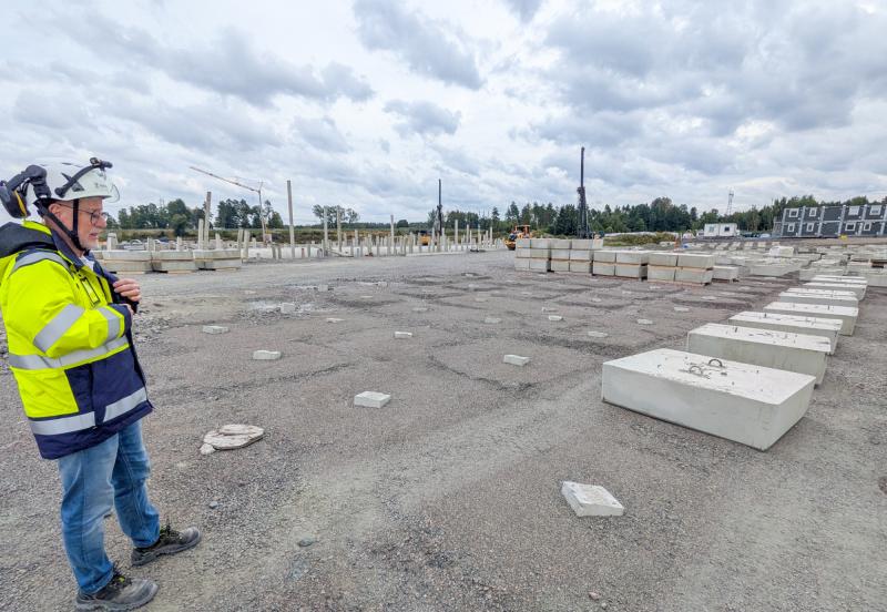En person i en varselväst och hjälm står på en byggarbetsplats med betongblock på en grusbelagd yta under en molnig himmel.