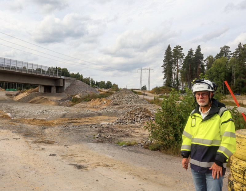 En man i en reflexväst och hjälm står på en byggarbetsplats nära en bro, med högar av grus och jord i bakgrunden.