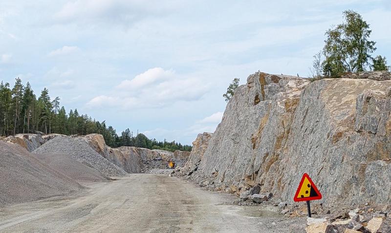En bred grusväg slingrar sig mellan höga klippväggar, och på höger sida syns en varningsskylt för jordskred. Träd kantar området under en molnig himmel.