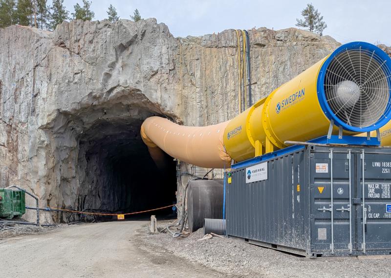 En stor tunnelingång i en klippig sluttning med en gul ventilationskanal ansluten till industriutrustning bredvid öppningen vid en grusväg.