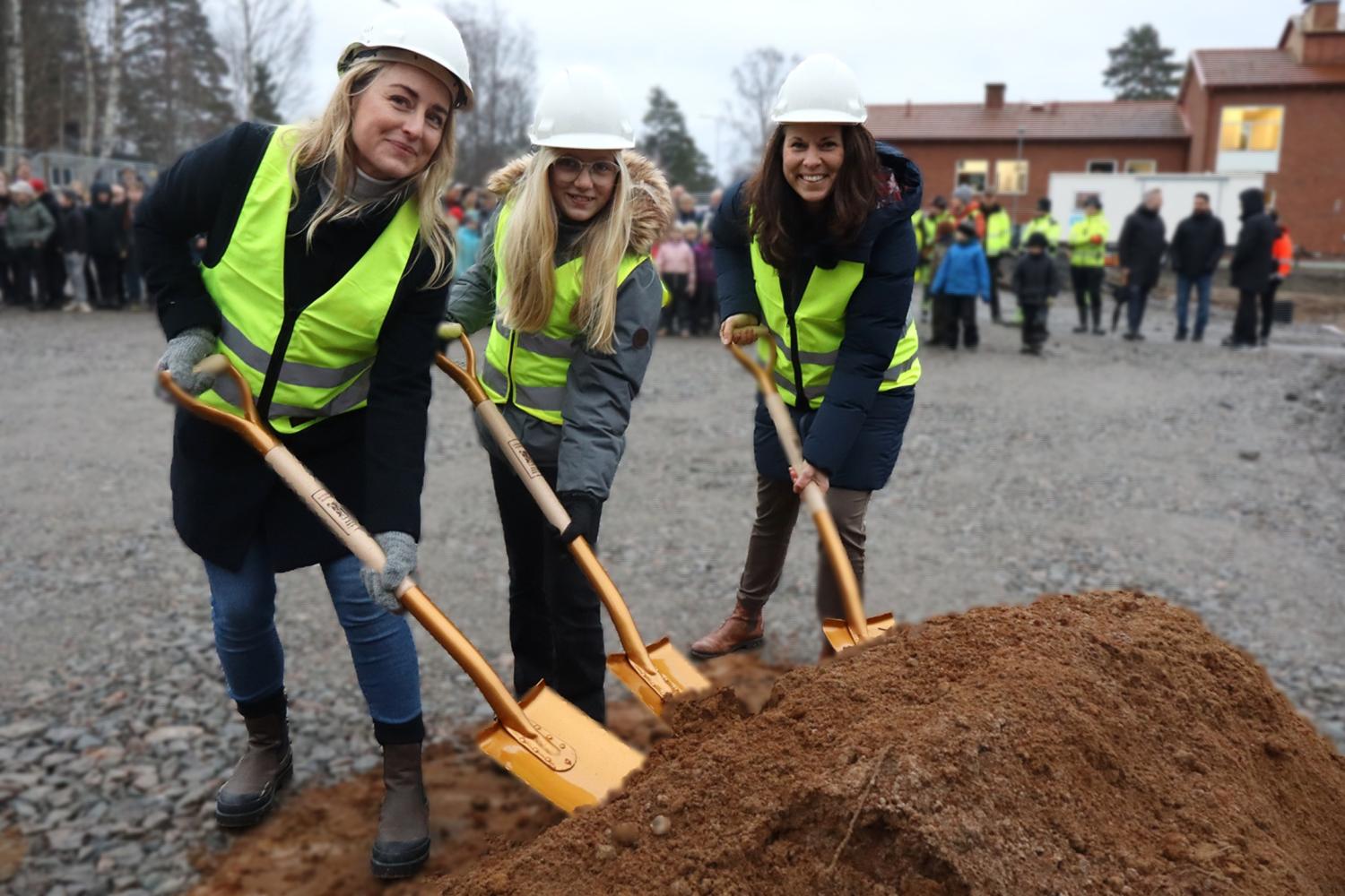 Tre kvinnor i skyddsvästar och hjälmar håller i spadar och tar första spadtaget på en byggarbetsplats, med en folkmassa och byggnader i bakgrunden.