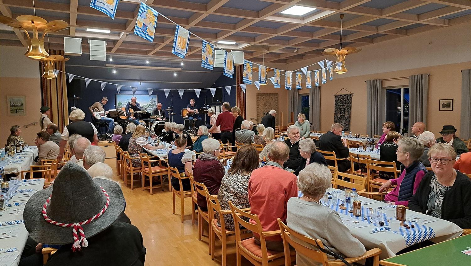 En grupp människor sitter vid långbord i en dekorerad hall och ser ett band uppträda på scenen. Blå och vita banderoller hänger från taket.