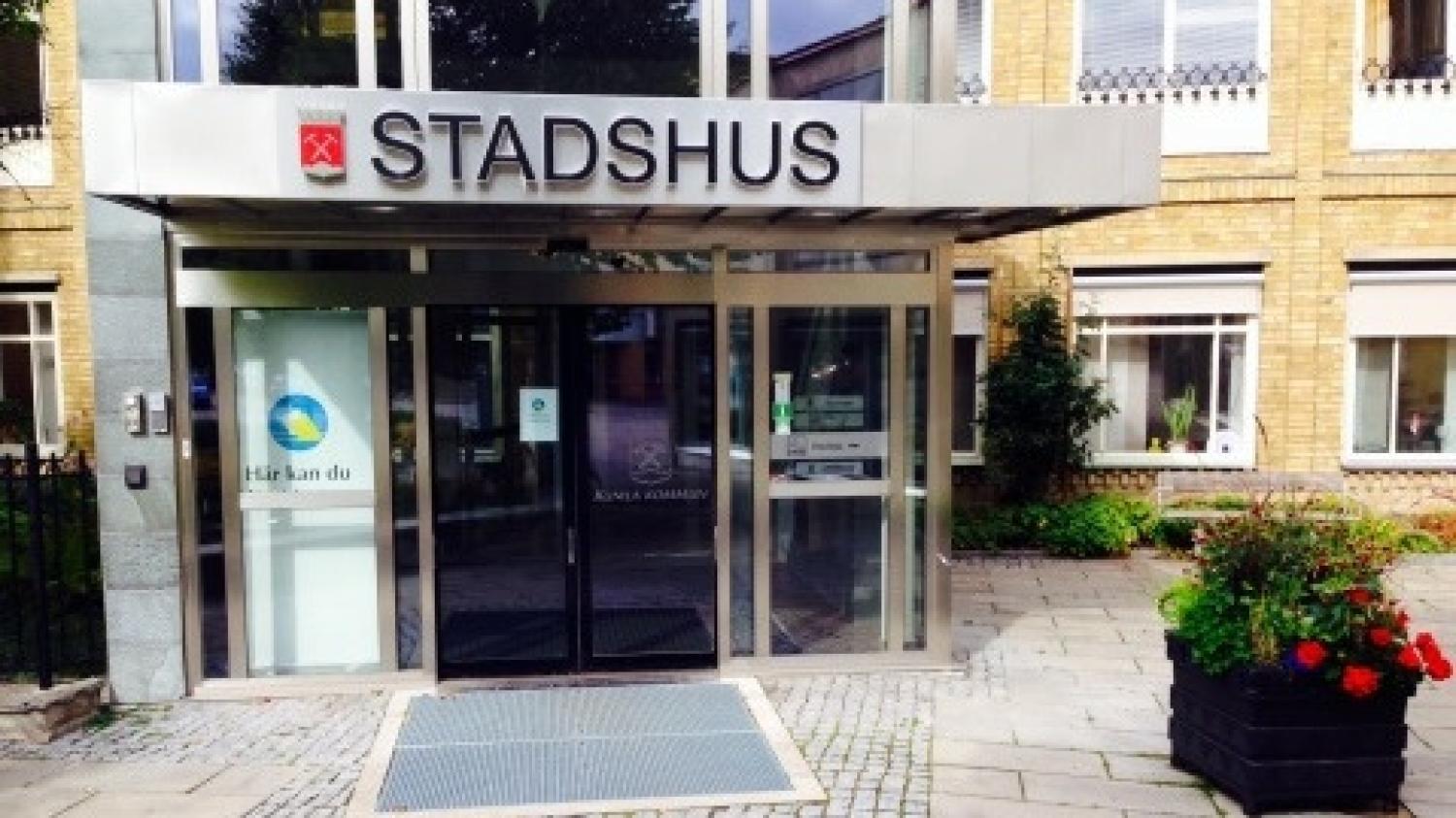 Ingången till en byggnad med namnet ”Stadshus”, med glasdörrar, en blomlåda och skyltar i fönstren; fasaden är av gula tegelstenar.