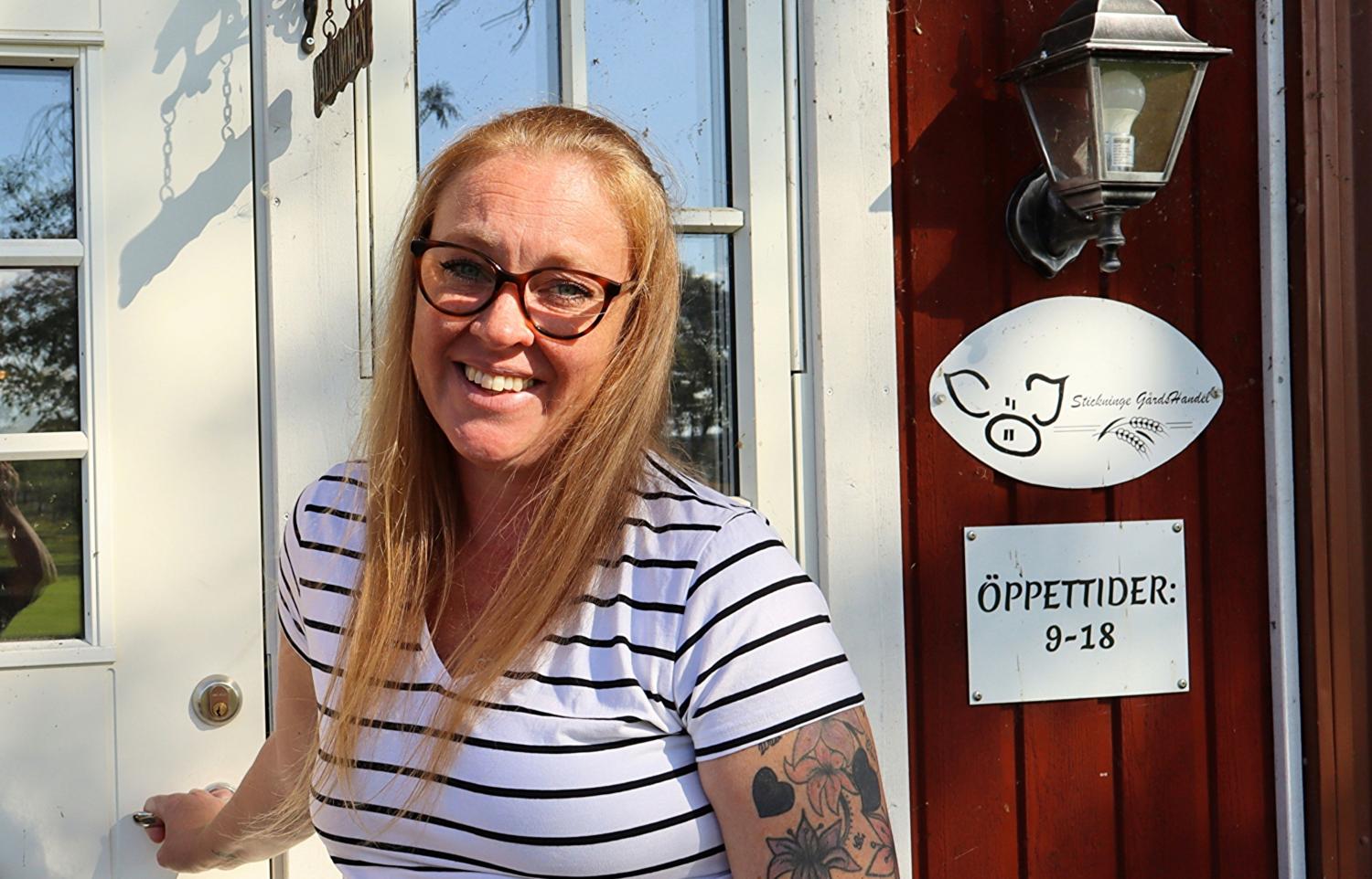 En kvinna med långt blont hår och glasögon, klädd i en randig skjorta, står leende i en dörröppning bredvid en skylt med öppettider.