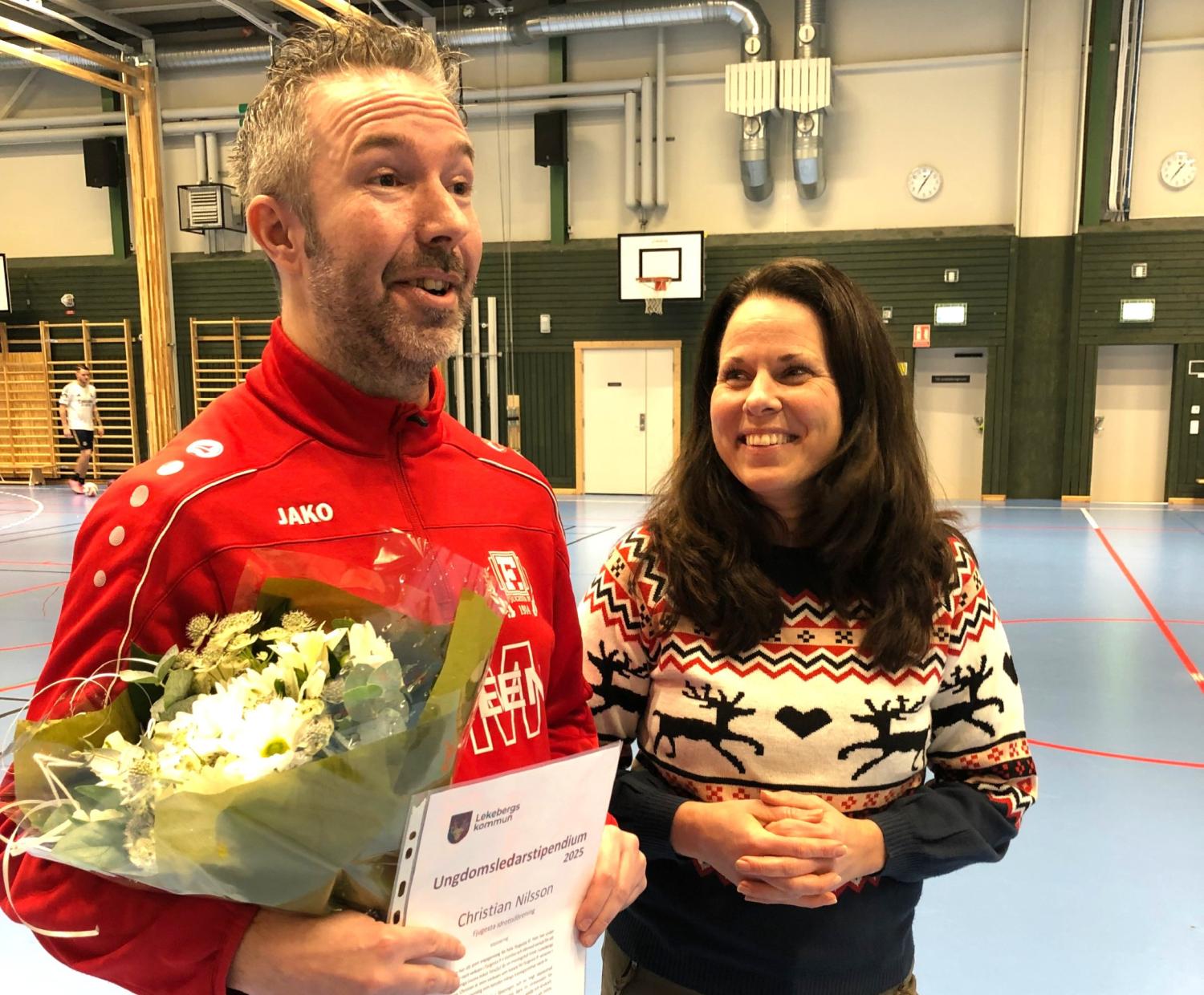 En man i röd sportjacka håller i en bukett och ett diplom och står bredvid en leende kvinna i en tröja med vintertema i en gymnastiksal.