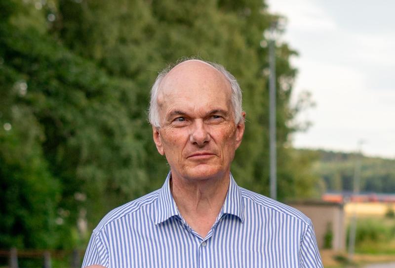 En äldre man med tunnnande grått hår och en randig skjorta står utomhus, med träd och en suddig byggnad i bakgrunden.