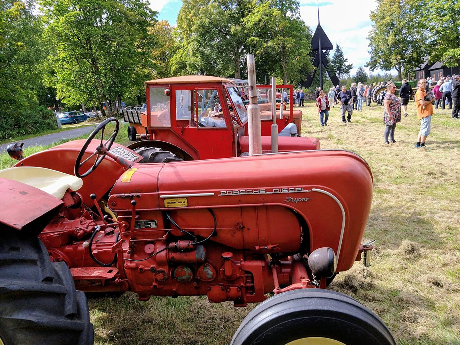 Två röda vintagetraktorer står parkerade på gräset vid ett utomhusevenemang, med människor samlade och träd i bakgrunden.