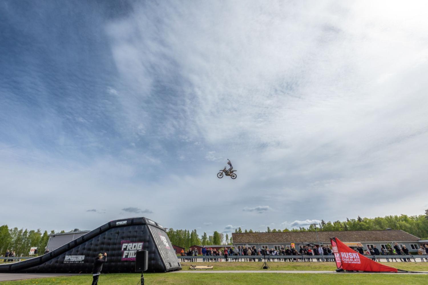 En motorcyklist gör ett hopp i luften över en ramp i ett utomhusevenemangsområde med åskådare och molnig himmel.