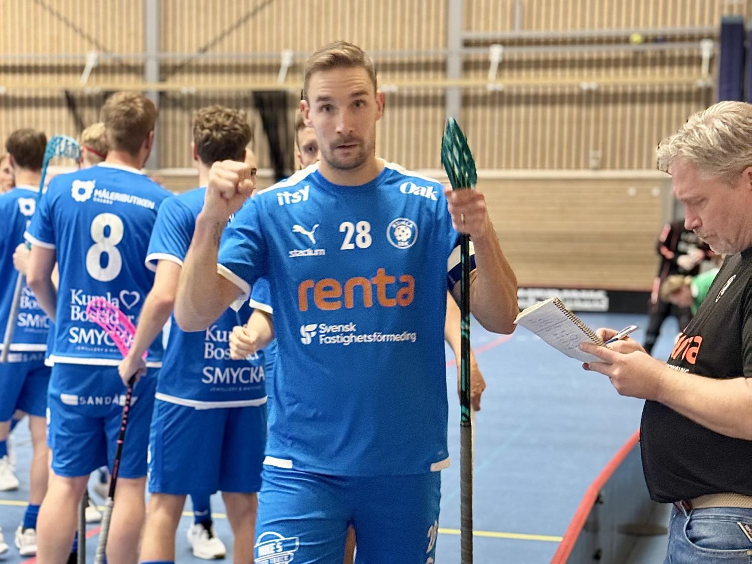 En manlig innebandyspelare i blå tröja med nummer 28 står och håller i en klubba, medan andra spelare och en man med ett anteckningsblock befinner sig i närheten inomhus.