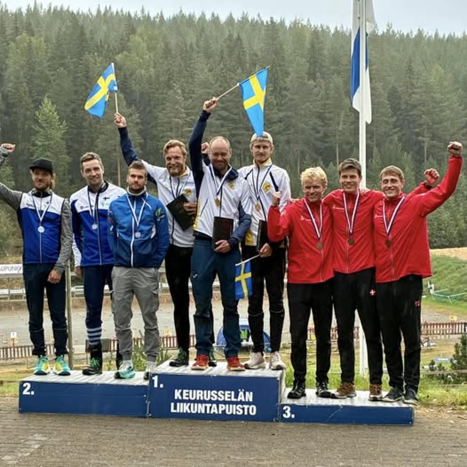 Tre lag står på ett vinnarpodium utomhus, med medaljer och flaggor i händerna, med första, andra och tredje plats markerade. Skog i bakgrunden.