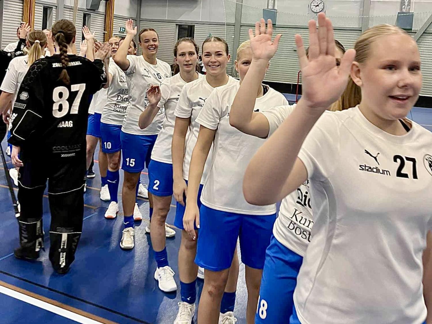 En grupp unga kvinnliga idrottare i vita tröjor och blå shorts ställer upp sig för att high fivea ett annat lag efter en match i en inomhushall.