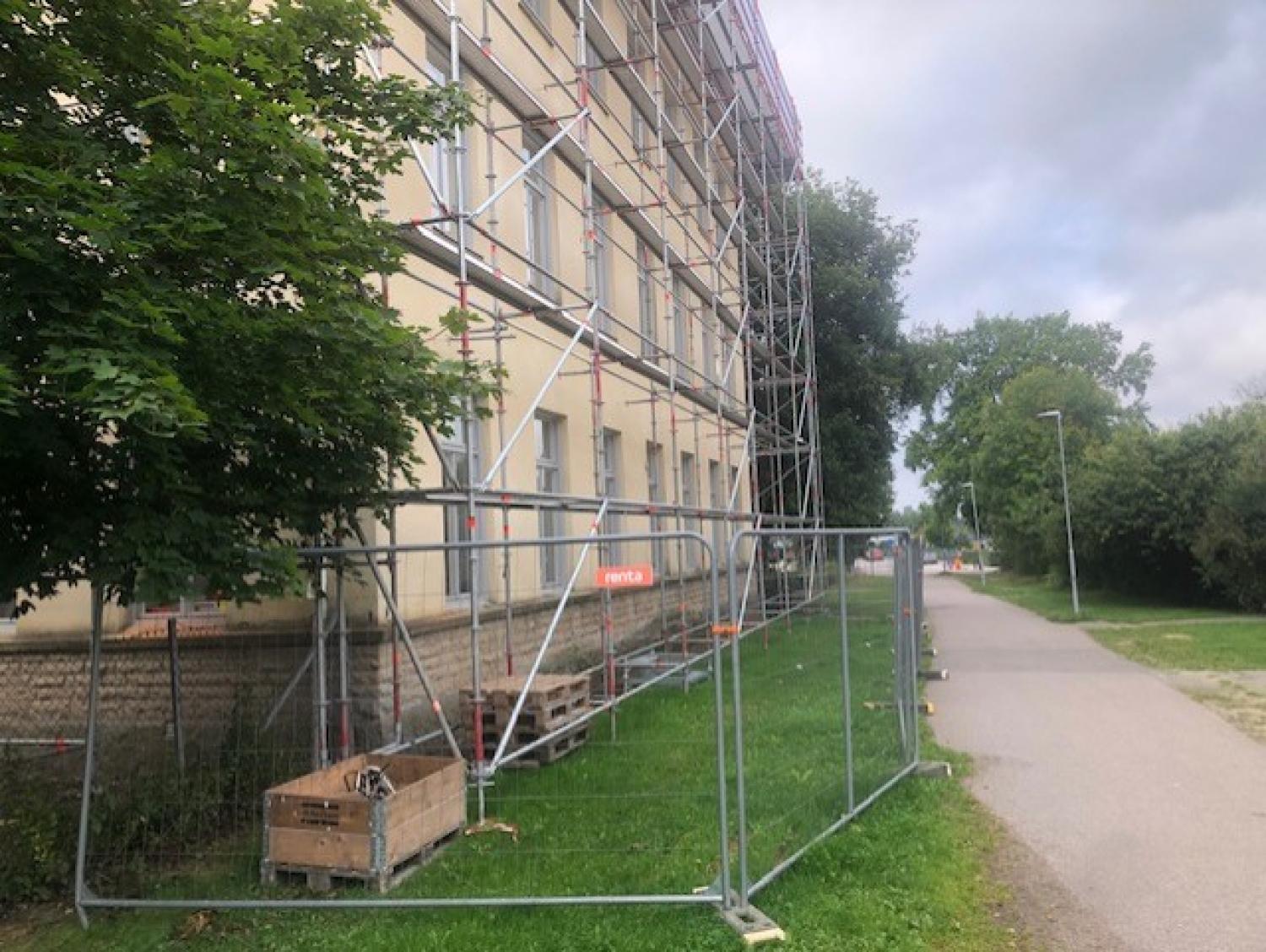 En byggnad under uppförande är omgiven av byggnadsställningar och ett tillfälligt metallstaket, med lastpallar och byggmaterial på gräset i närheten. En asfalterad stig löper längs med platsen.
