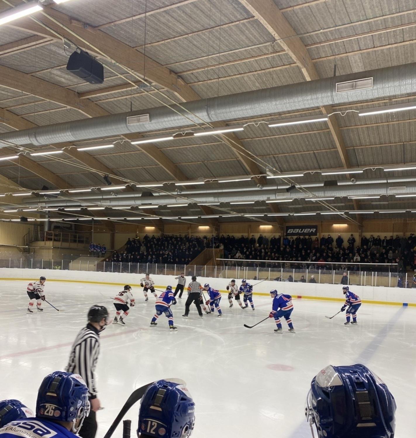 Två ishockeylag möts i mitten av isen på en inomhusrink, med domare i närheten och en mängd åskådare som tittar på från läktaren.