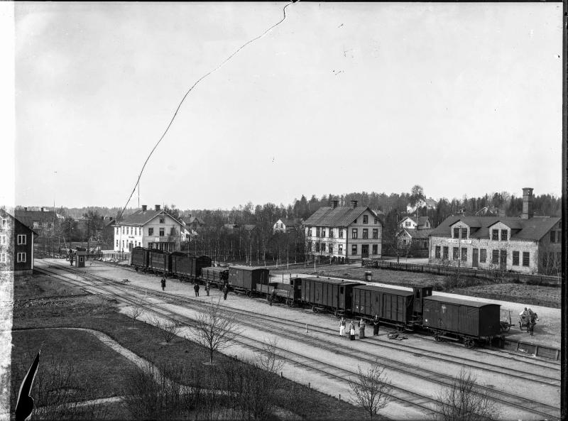 Svartvitt foto av en liten järnvägsstation med flera godsvagnar på spåren, några personer som står i närheten samt byggnader och träd i bakgrunden.