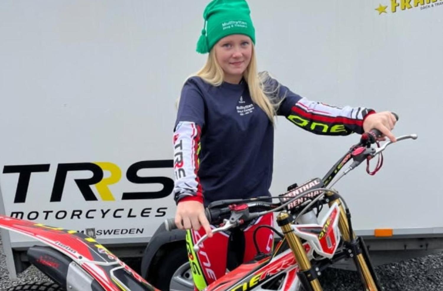 Person i en grön mössa som står vid en motorcykel, framför en TRS Motorcycles trailer.