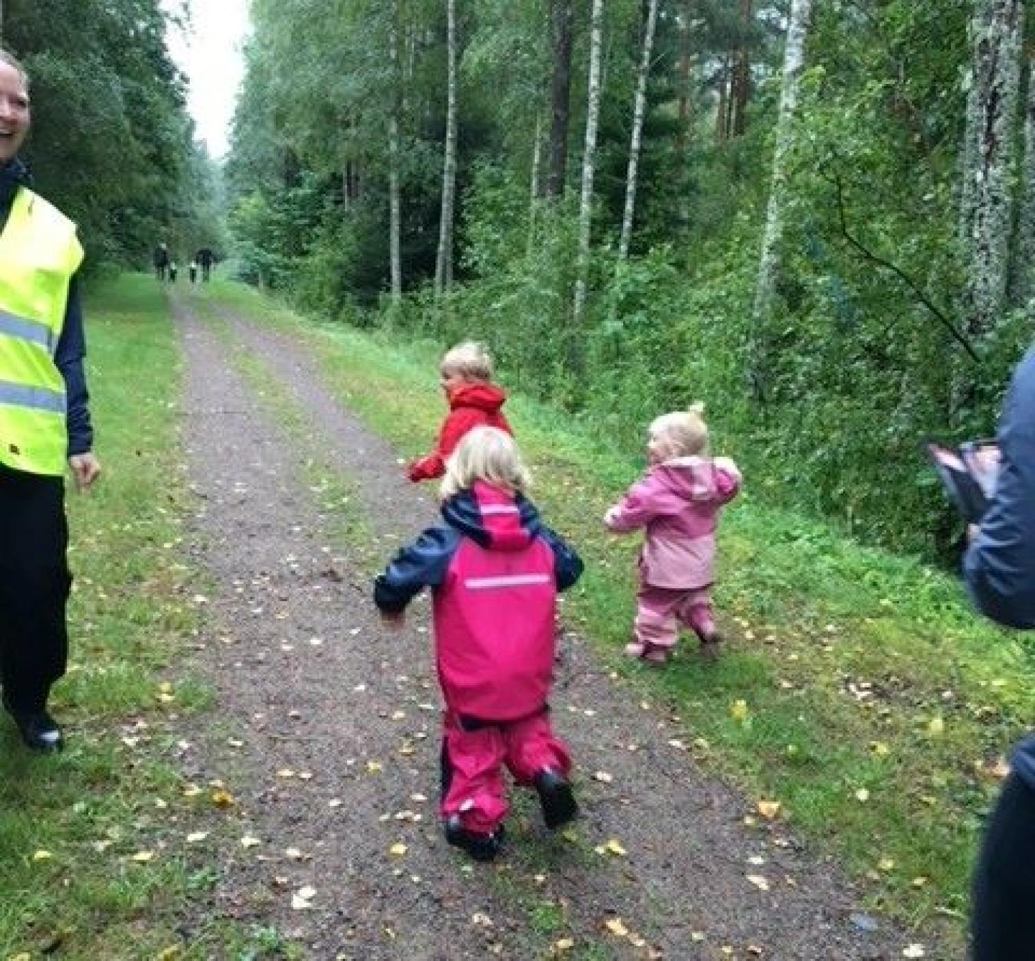 Tre barn i färgglada friluftskläder går på en skogsstig i sällskap av vuxna. Träd och grönska omger leden.