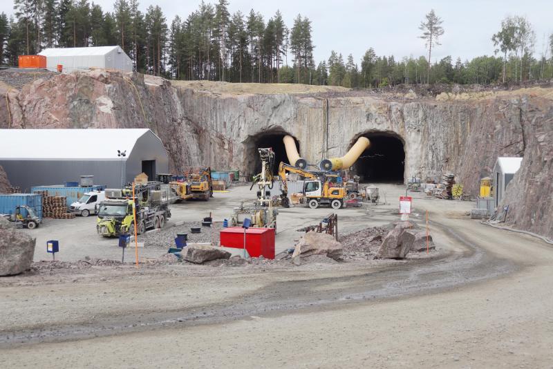 En byggarbetsplats med tunga maskiner framför två stora tunnelingångar i en klippig sluttning, omgiven av skog och tillfälliga byggnader.
