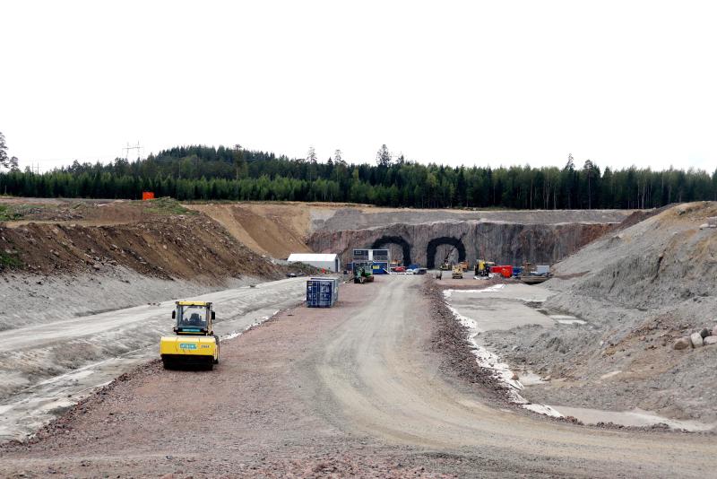 En byggarbetsplats med tunga maskiner och utrustning framför två tunnelingångar, omgiven av jordhögar och en skog i bakgrunden.
