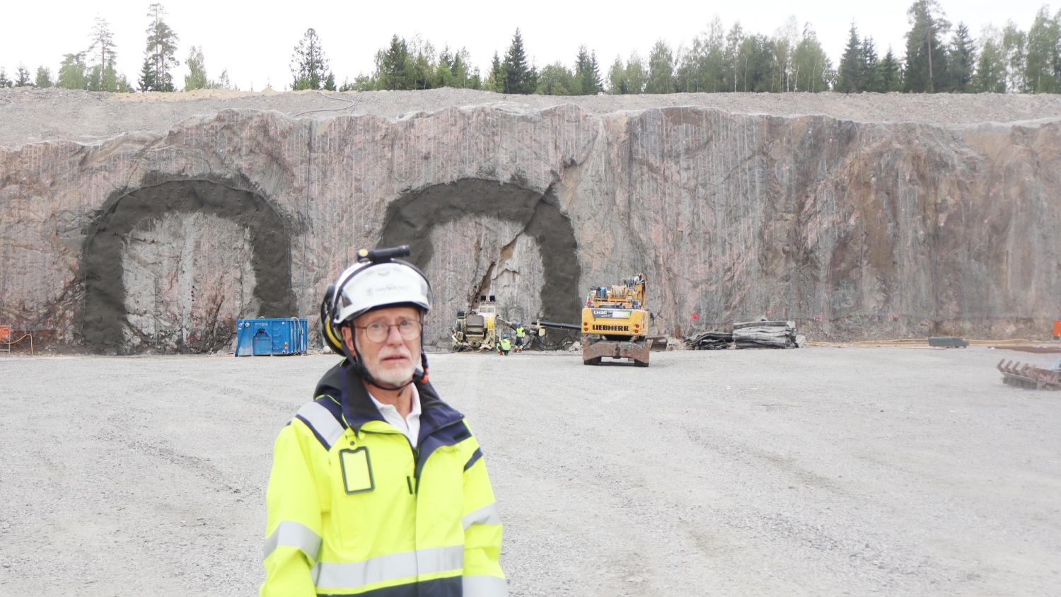 En man i en reflexväst och hjälm står framför två tunnelingångar som huggits ut i en klippig sluttning, med byggmaskiner och arbetare synliga i bakgrunden.