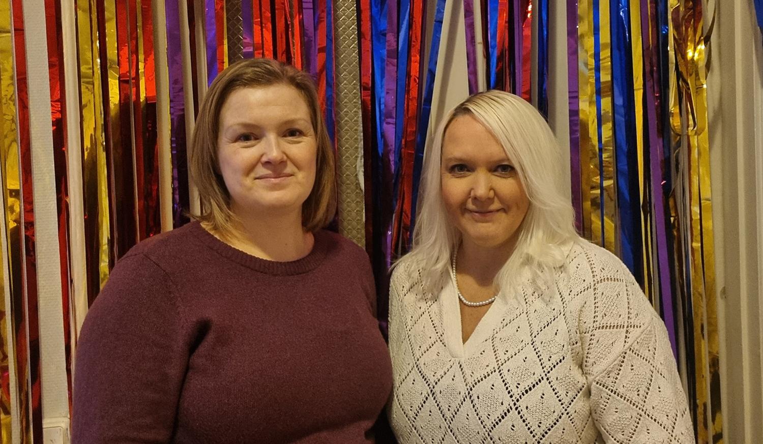 Två kvinnor står sida vid sida framför färgglada metalliska serpentiner, den ena bär en rödbrun tröja och den andra en vit tröja.
