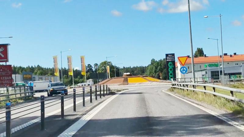 En väg med metallräcken leder till en rondell med en gul mittö. Bilar står parkerade till vänster, och på båda sidor syns byggnader och skyltar. Himlen är klar och blå.
