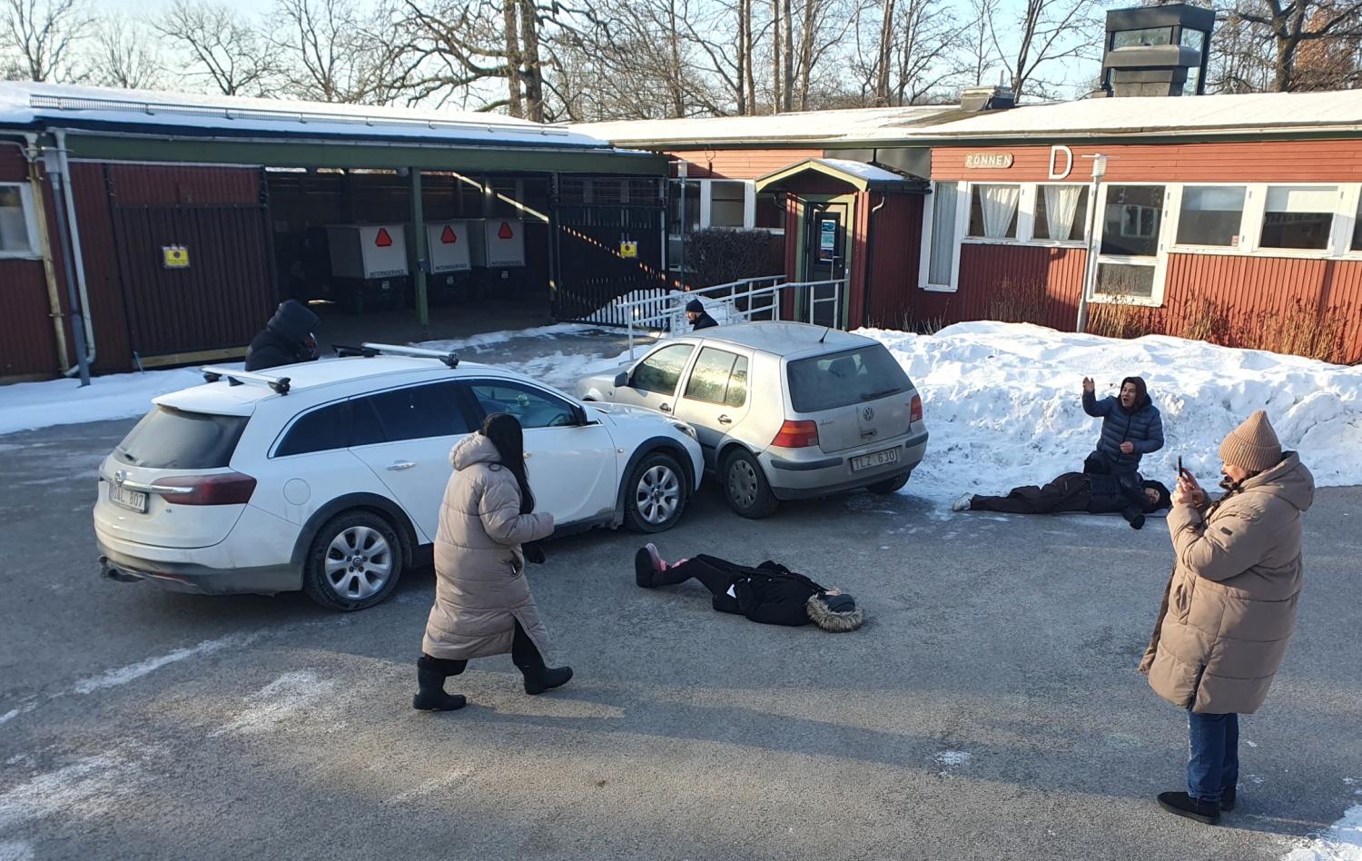 Flera personer i vinterkläder står eller ligger på en snöig parkeringsplats bredvid två parkerade bilar utanför en byggnad, några verkar posera eller ta bilder.