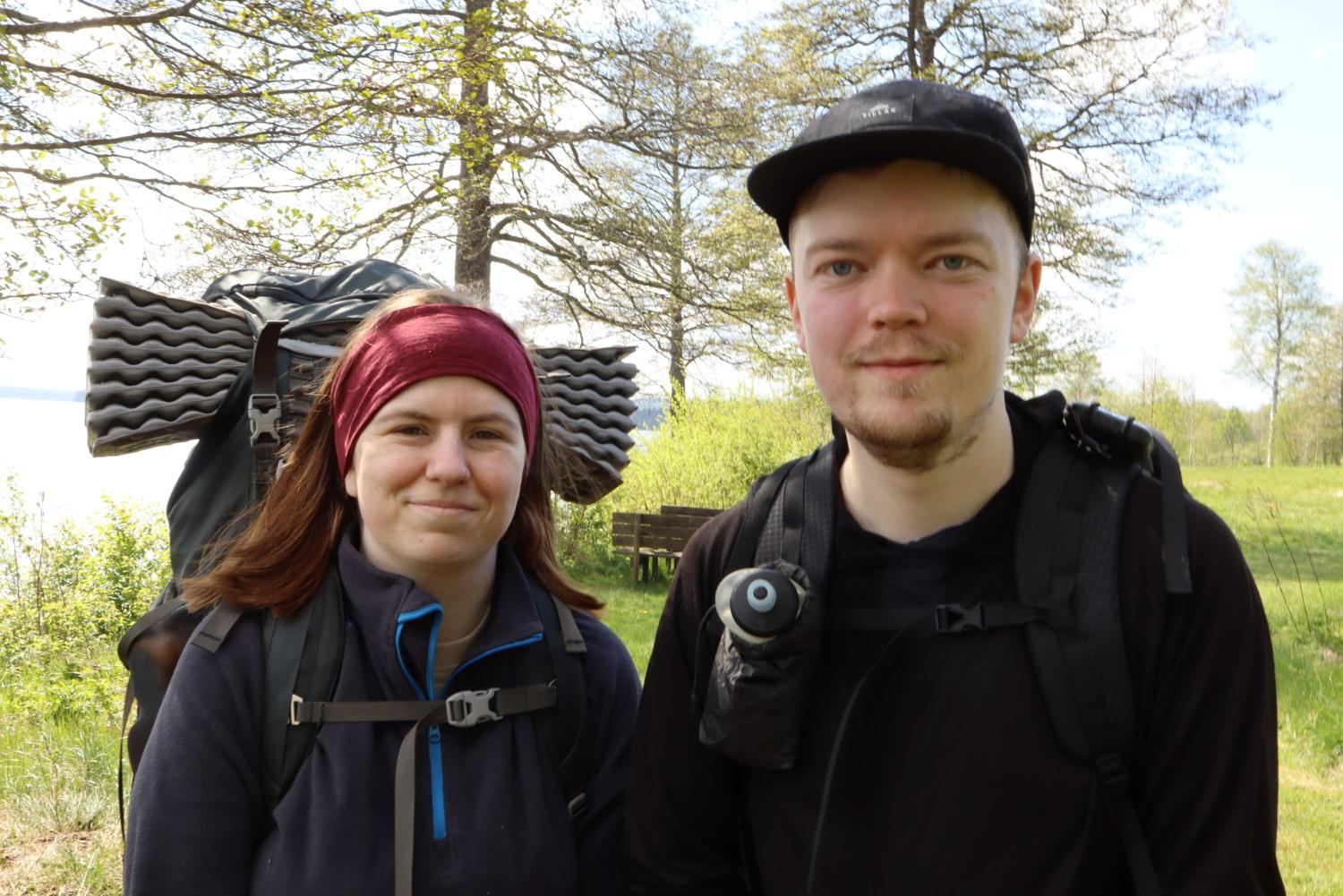 Två personer med ryggsäckar och friluftsutrustning står utomhus i ett gräsbevuxet skogsområde med träd och en sjö i bakgrunden.
