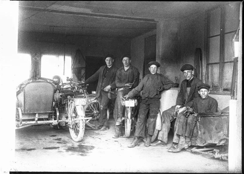 Fem män i arbetskläder och kepsar står eller sitter i ett garage bredvid två veteranmotorcyklar, varav en med sidovagn, omgivna av verkstadsverktyg och utrustning.