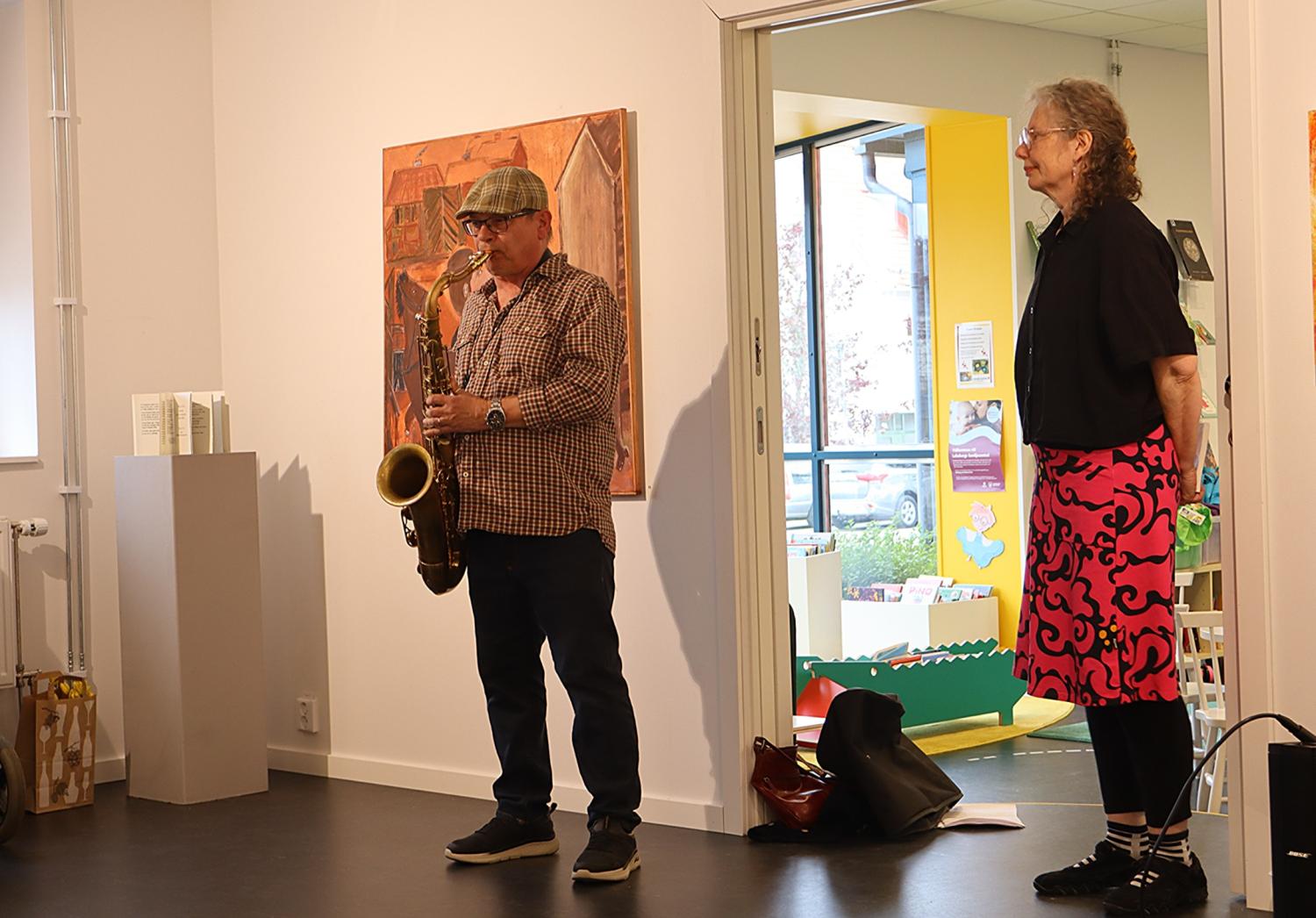 En man spelar saxofon medan en kvinna står bredvid i ett konstgalleri, med konstverk på väggarna och en öppen dörr som visar ett färgglatt rum bortom.