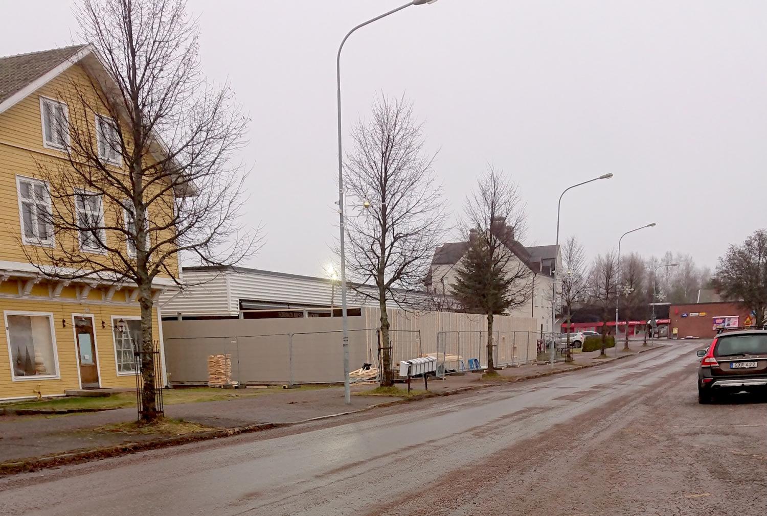 Gatuvy en molnig dag med en gul byggnad till vänster och byggarbetsplats i bakgrunden, flankerad av bladlösa träd och lyktstolpar.