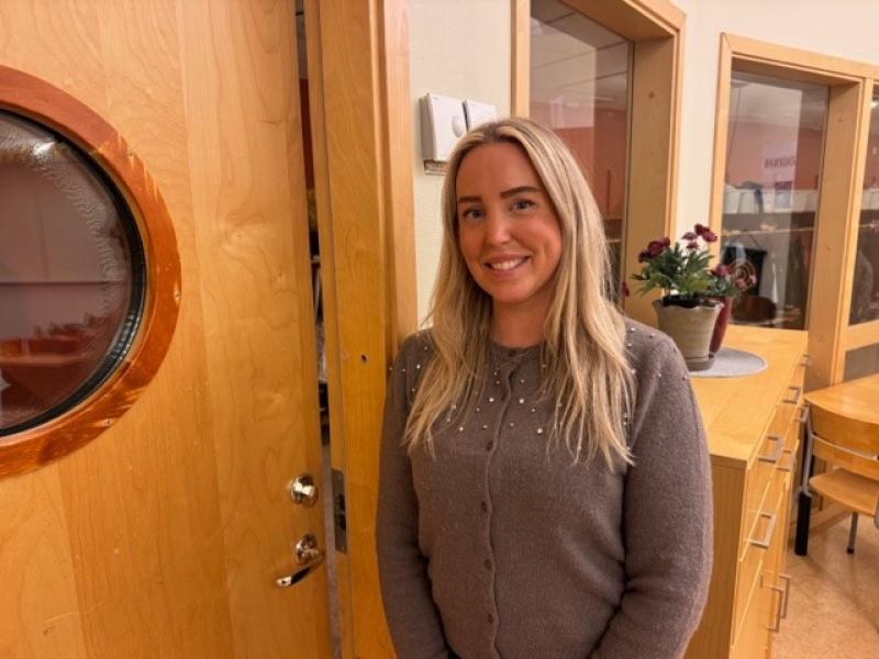 En kvinna med långt blont hår och grå tröja står inomhus vid en trädörr, med skåp, stolar och en krukväxt i bakgrunden.