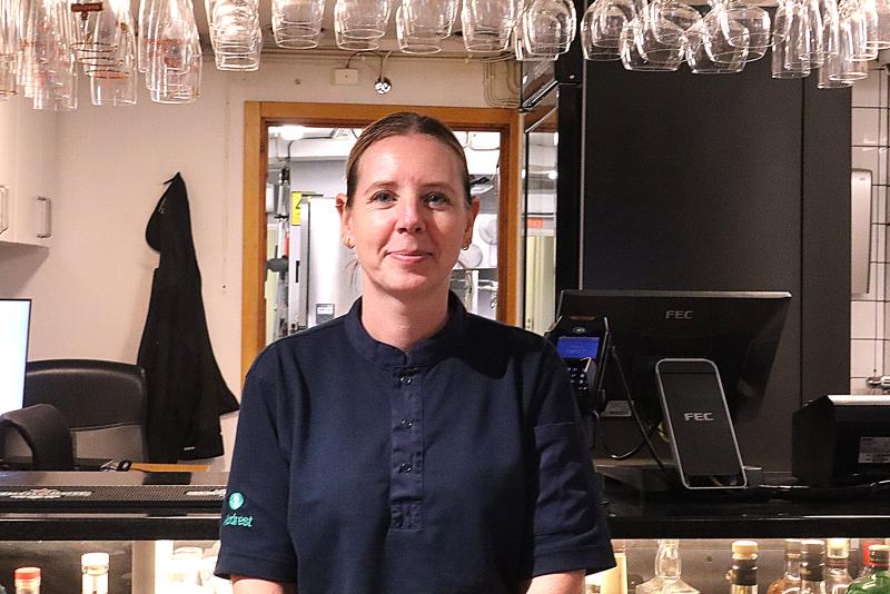 En kvinna i mörk skjorta står bakom en bardisk med glasvaror hängande ovanför och flaskor synliga nedanför.
