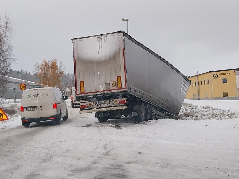 Ett stort lastbilssläp har glidit av en snöig väg ner i ett dike bredvid en byggnad, medan en vit skåpbil från NRC Group står parkerad i närheten.