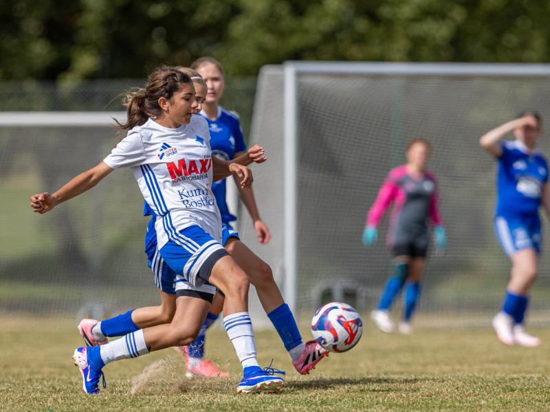 En fotbollsspelare i en vit och blå tröja sparkar bollen under en match, med andra spelare och ett mål synliga i bakgrunden.