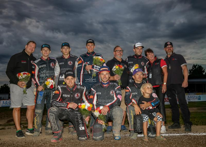 En grupp speedwayförare i tävlingsdräkter och teamets personal poserar tillsammans utomhus, varav några håller i blomsterbuketter, med en molnig himmel i bakgrunden.